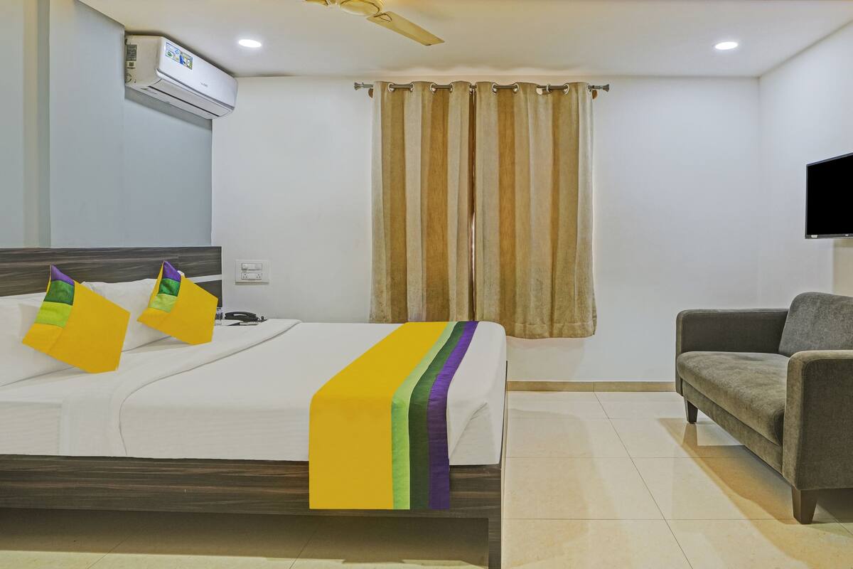A hotel room with a bed, a sofa, a TV mounted on the wall, and a window with curtains.