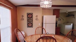 Un coin repas comprenant une table en bois et quatre chaises, un réfrigérateur et une fenêtre avec des stores.