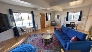 A spacious living room with a blue sofa, a round coffee table, and a colorful rug. There is a TV on the left, a chair, and a desk with a lamp. - Blue Skye: Downtown Getaway (Bishop)
