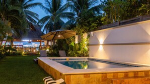 Piscina com deck revestido, rodeada por palmeiras e uma estrutura de telhado de palha.