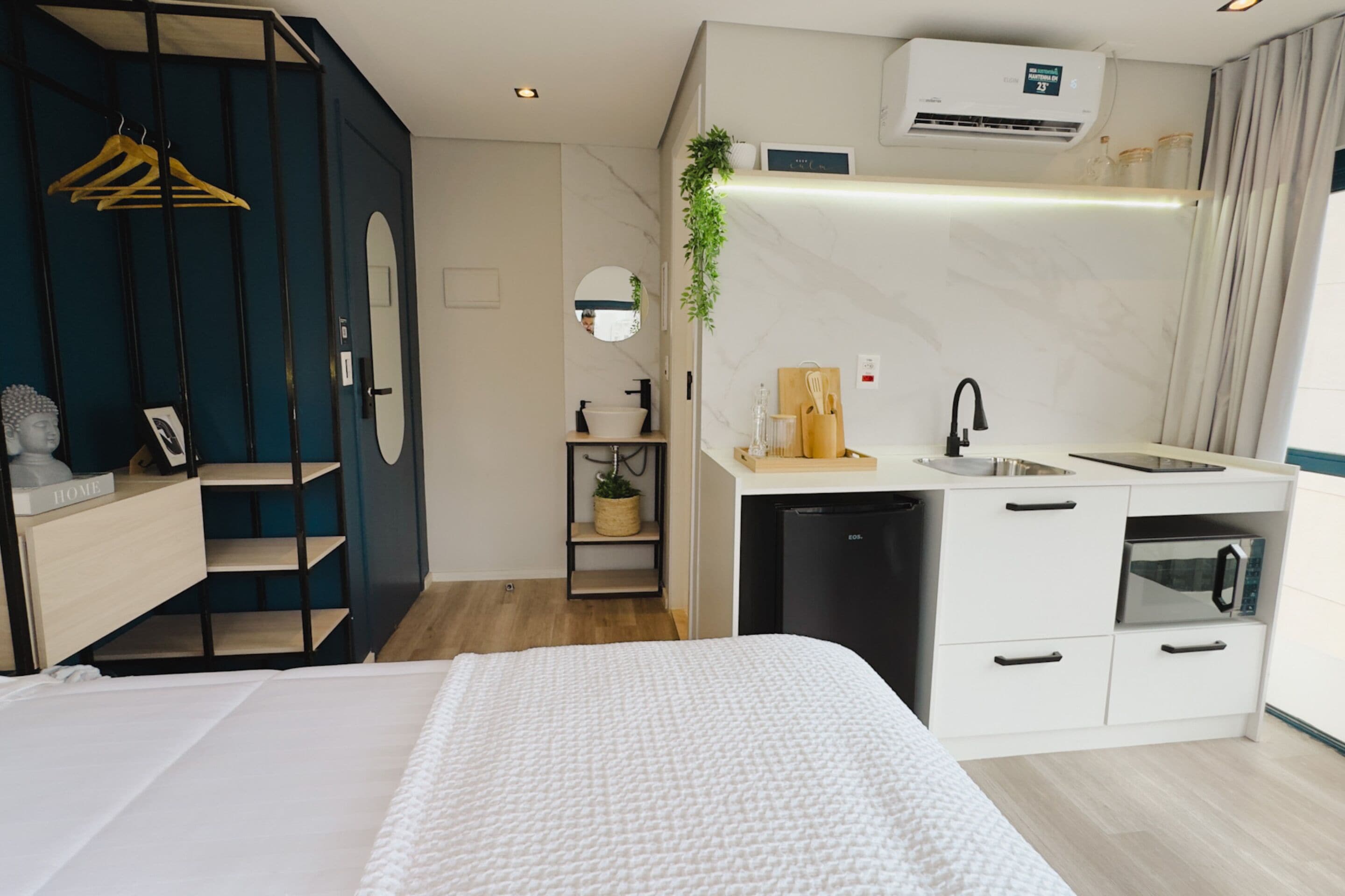 A modern hotel room with a bed, a compact kitchenette, a small bathroom, and a dark-colored wardrobe.