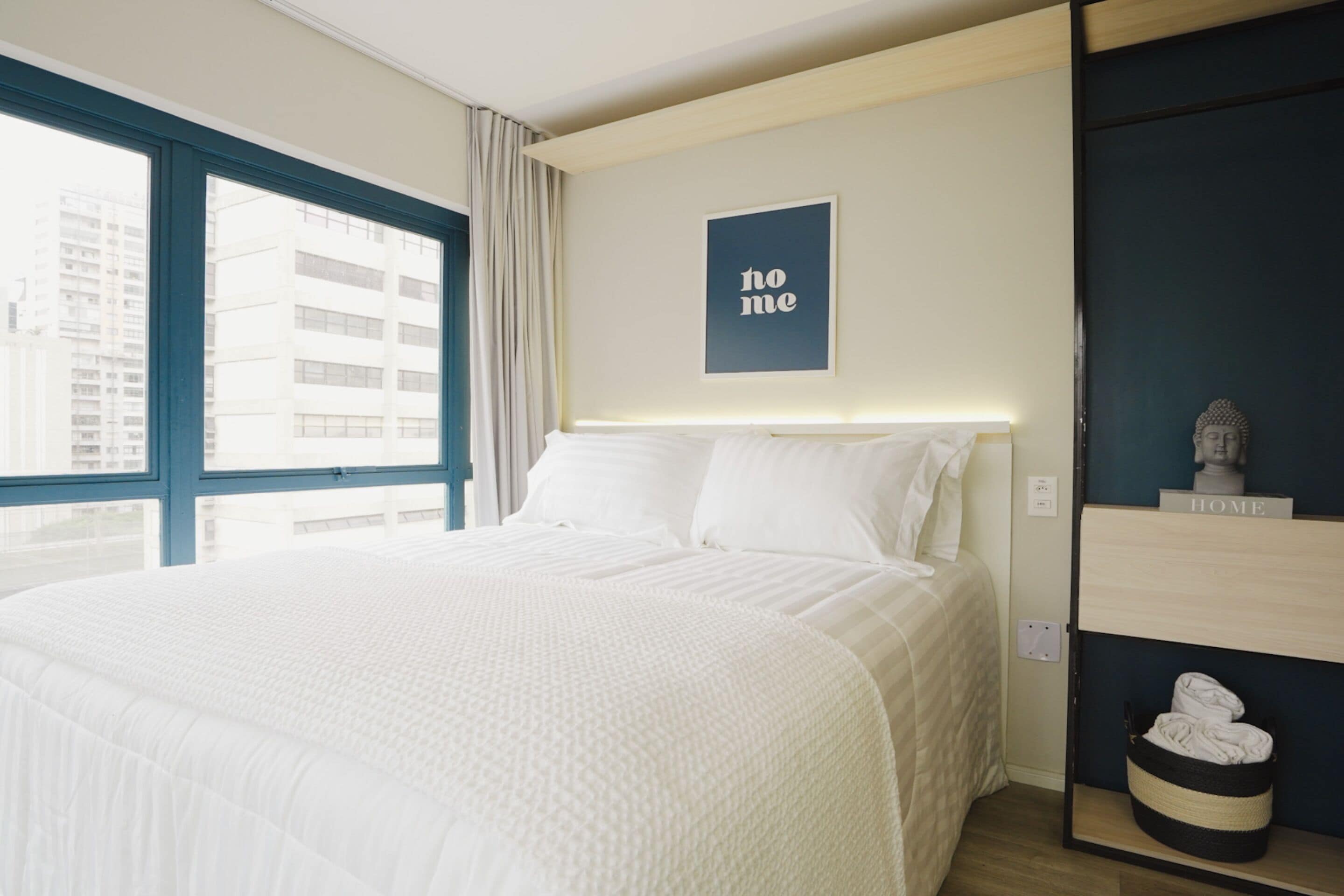 Ein Hotelzimmer mit einem großen Bett, Blick auf Gebäude durch ein Fenster, einem 'home'-Schild an der Wand und einer kleinen Ablage mit einer Statue.