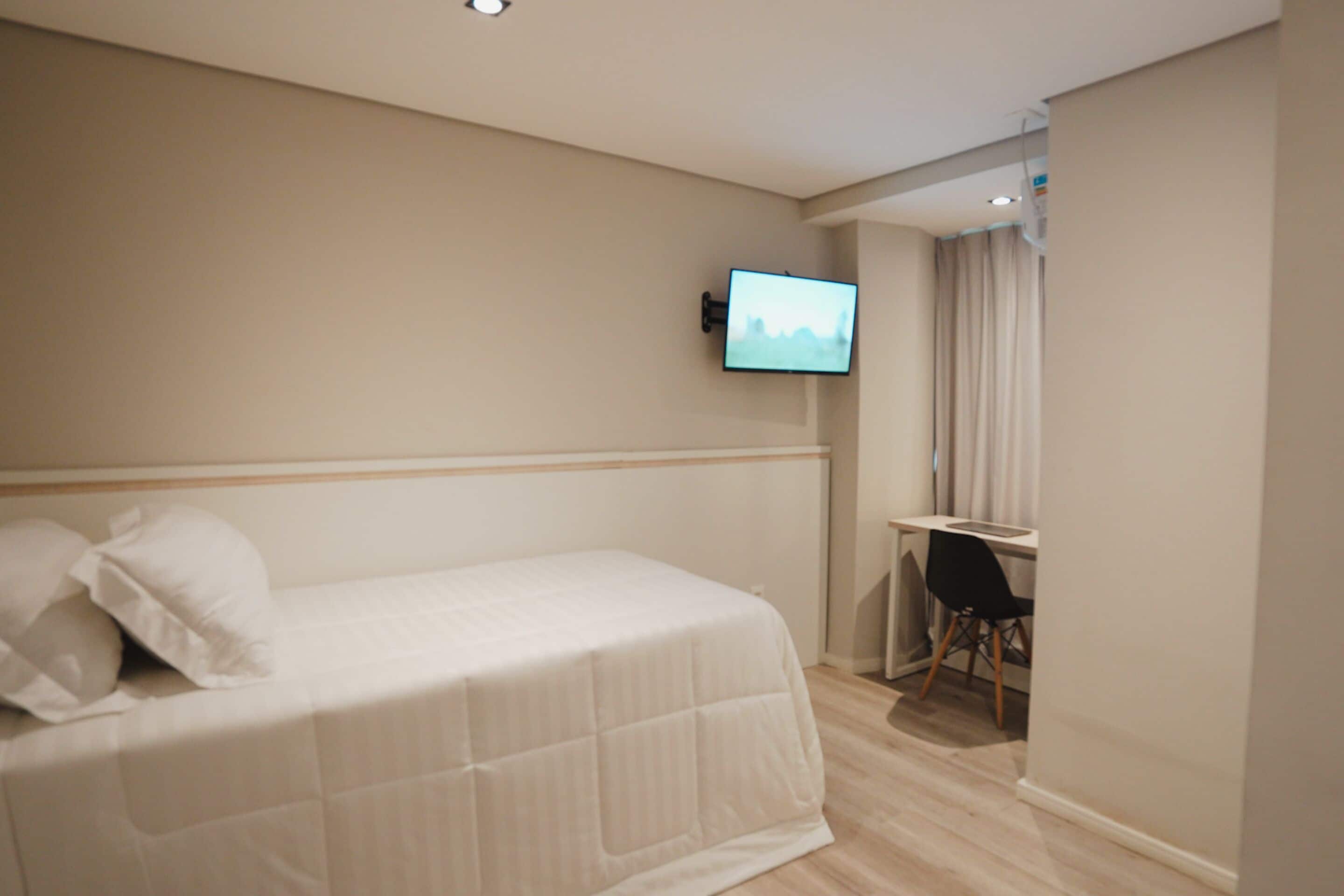 A hotel room with a bed, a desk, a chair, and a television mounted on the wall.