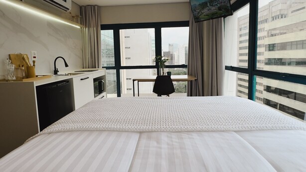 A hotel room with a large bed, a desk, and a view of the cityscape through large windows. - Apê Bela Cintra (São Paulo)