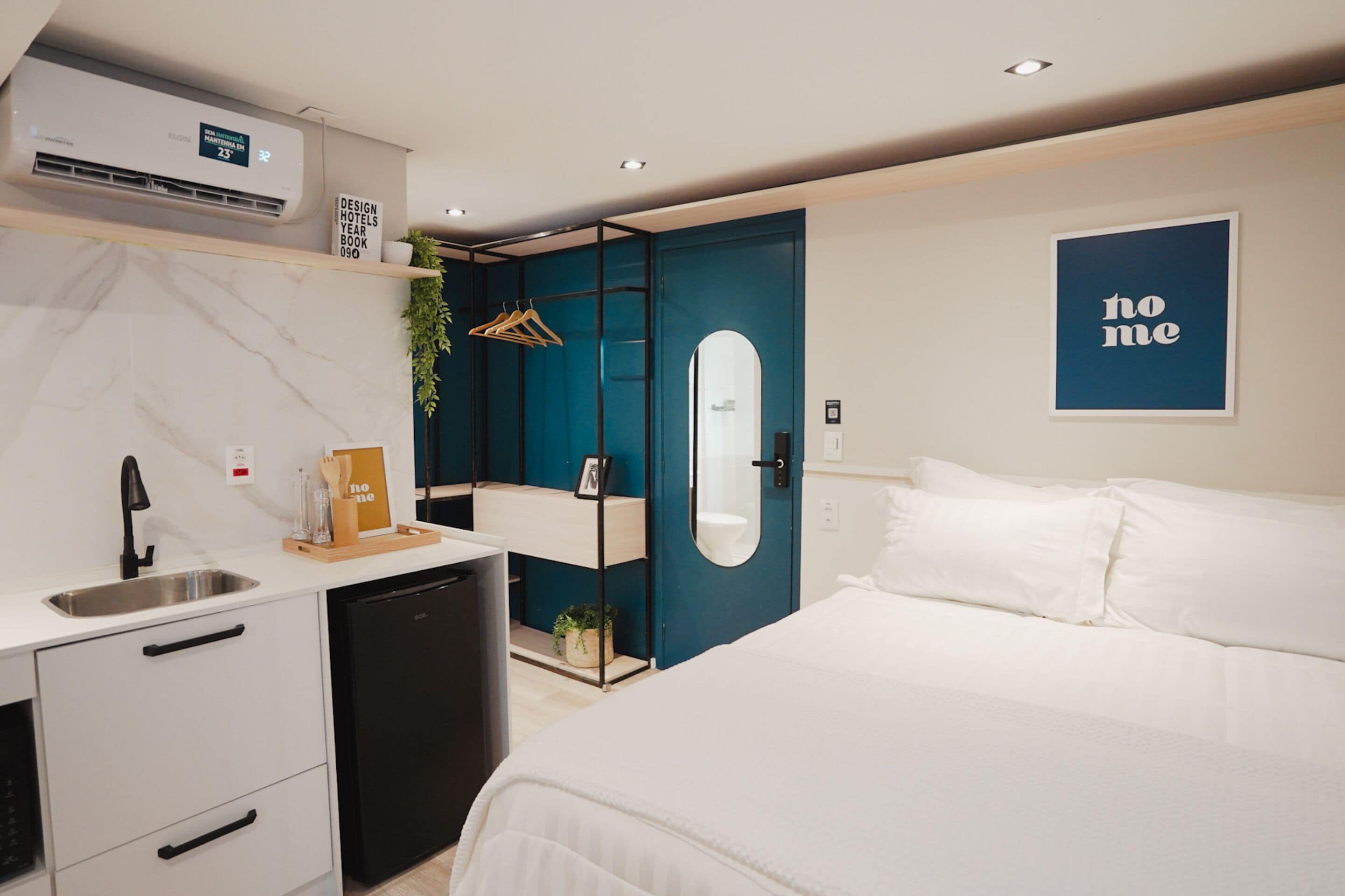 A modern hotel room with a bed, a small kitchenette, a mirror, and a blue door.
