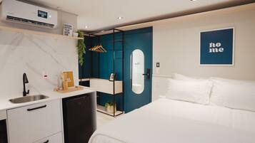 A modern hotel room with a bed, a small kitchenette, a mirror, and a blue door.