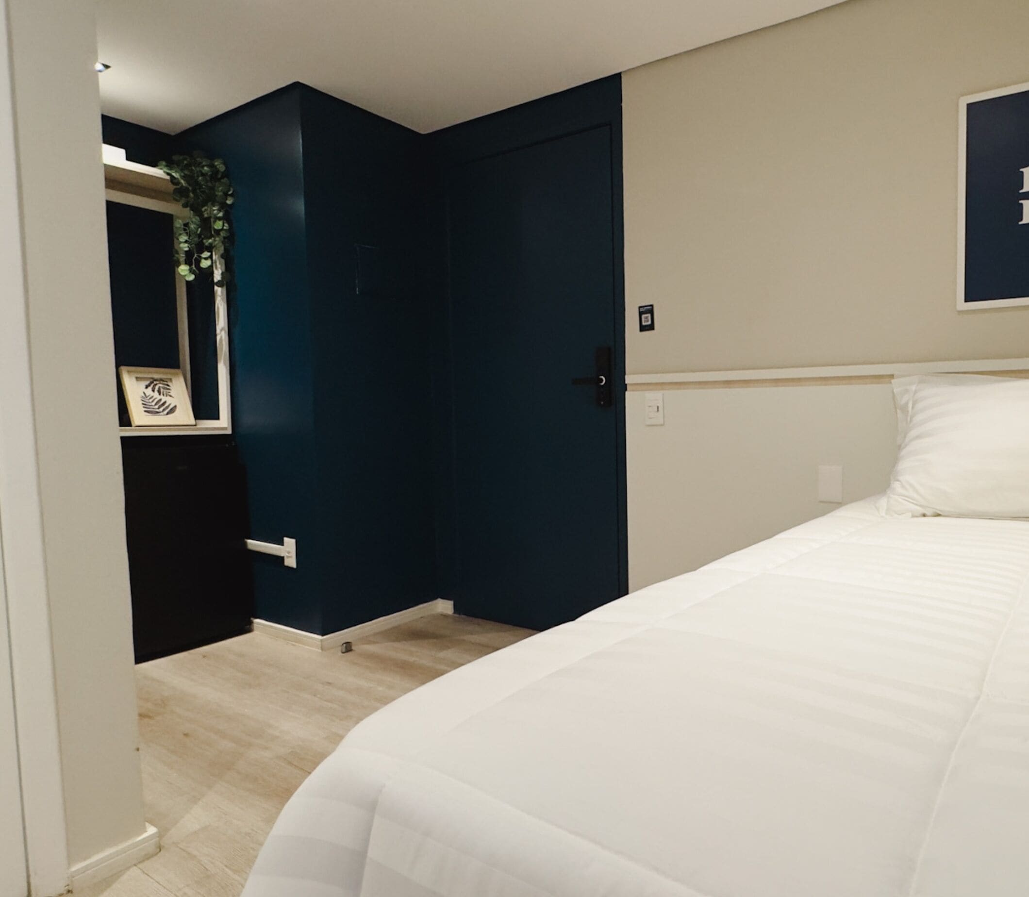 A bedroom with a bed, a dark blue door, a white wall, and a small plant on the left.