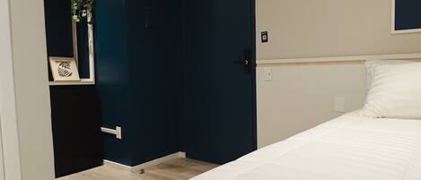 A bedroom with a bed, a dark blue door, a white wall, and a small plant on the left.