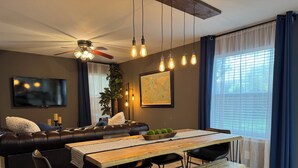 Ein modernes Wohnzimmer mit einem dunkel-ledernen Sofa, einem Holzespeisetisch mit Bänken, einem Deckenventilator und einem großen Fenster mit Vorhängen.
