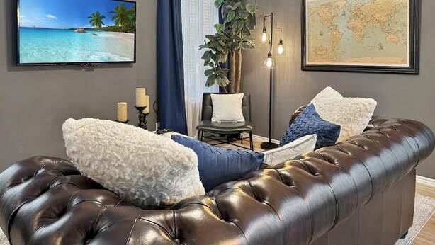 Eine dunkelbraune Ledersofa mit weißen und blauen Kissen, ein Flachbildfernseher, eine Weltkarte und eine Pflanze in einem Raum mit grauen Wänden und Holzböden.