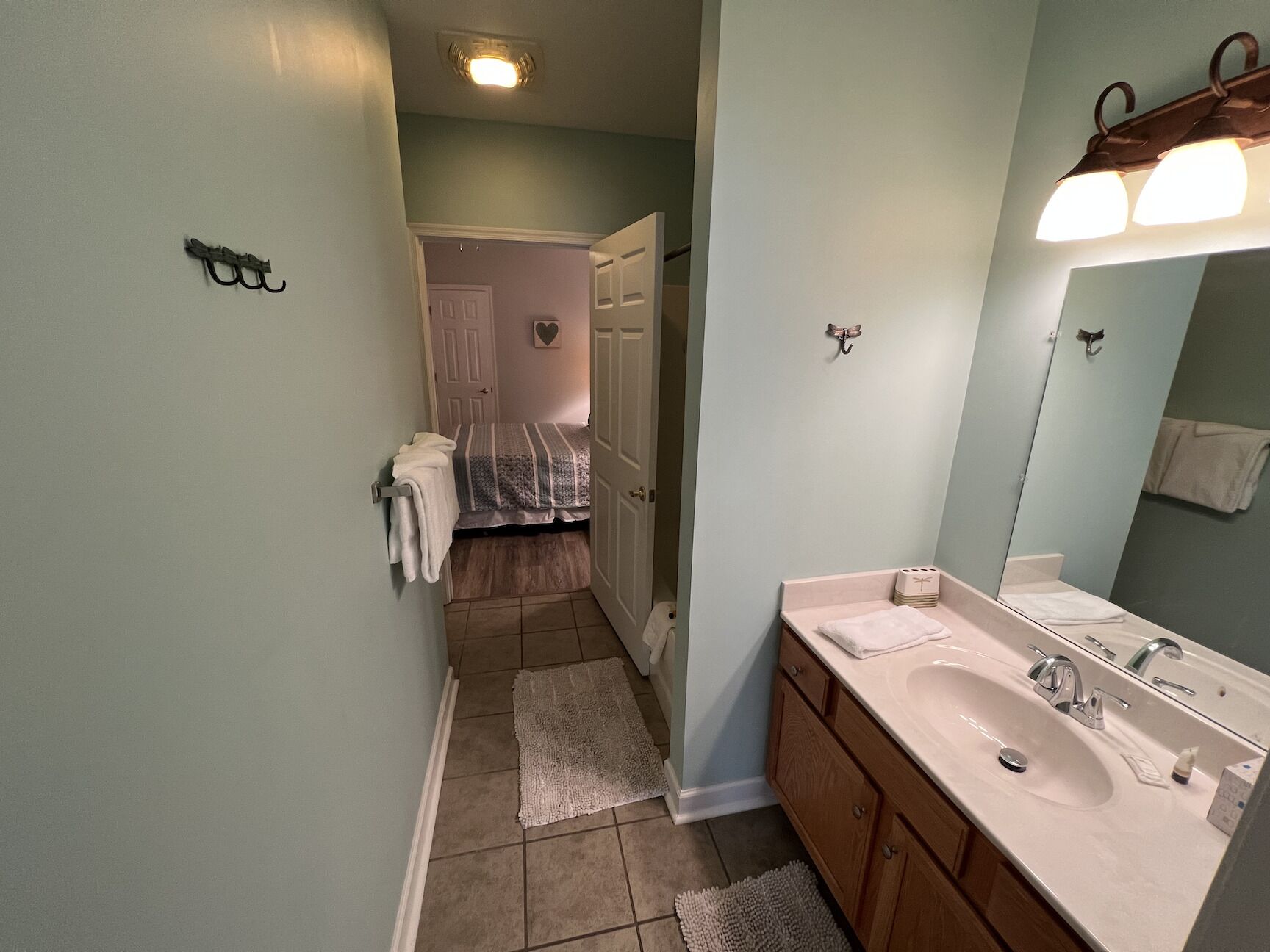 A bathroom with a sink, mirror, and towel rack. There is a door leading to a bedroom with a bed and a door.
