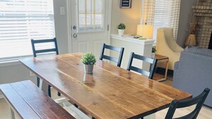 Un comedor con una mesa de madera, bancos y sillas negras. Se observa una puerta con ventana, una lámpara y una pequeña planta en maceta sobre una estantería.