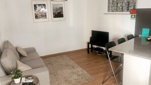A modern living room with a grey sofa, a small side table with a plant, a black TV stand, and a dining area with a black table and chairs.