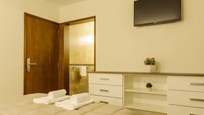 Ein Hotelzimmer mit Holztür, einem Bett mit weißer Bettwäsche, einem flachbildfernseher an der Wand und einer kleinen Pflanze auf dem Nachttisch.