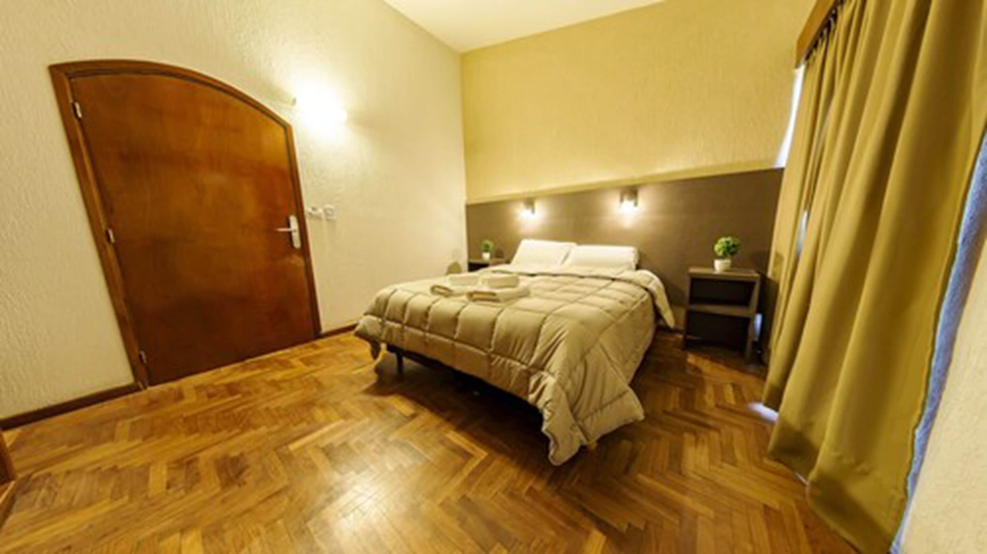 Une chambre avec un plancher en bois, un lit recouvert d’une couette beige, une table de chevet avec une lampe et une porte.