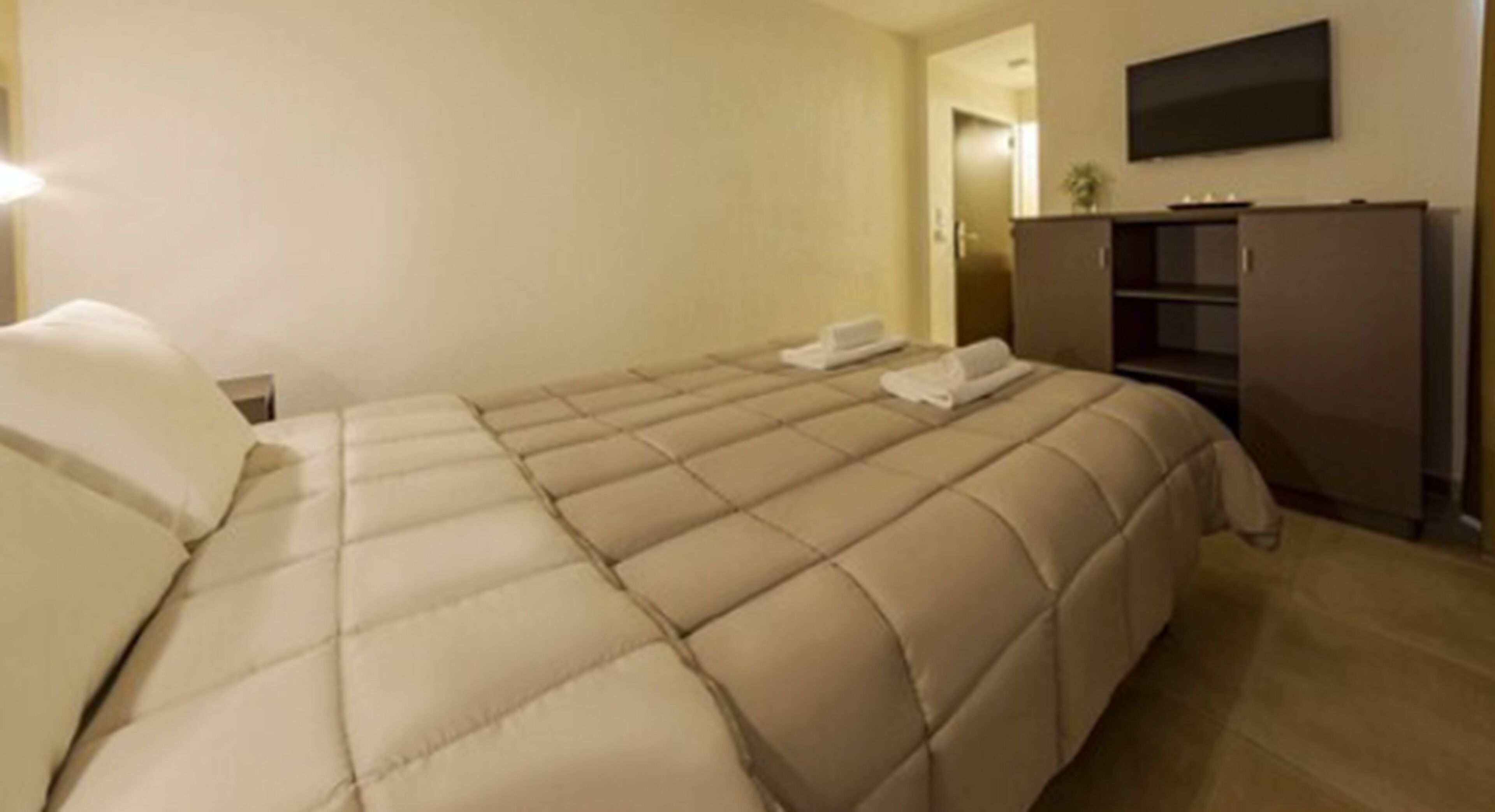 A hotel room with a large bed, a flat-screen TV mounted on the wall, and a minibar cabinet.
