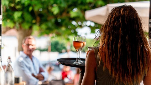 A person holding a tray with a glass of drink at an outdoor dining area with tables and chairs. - Auberge Koskenia (Bidart)