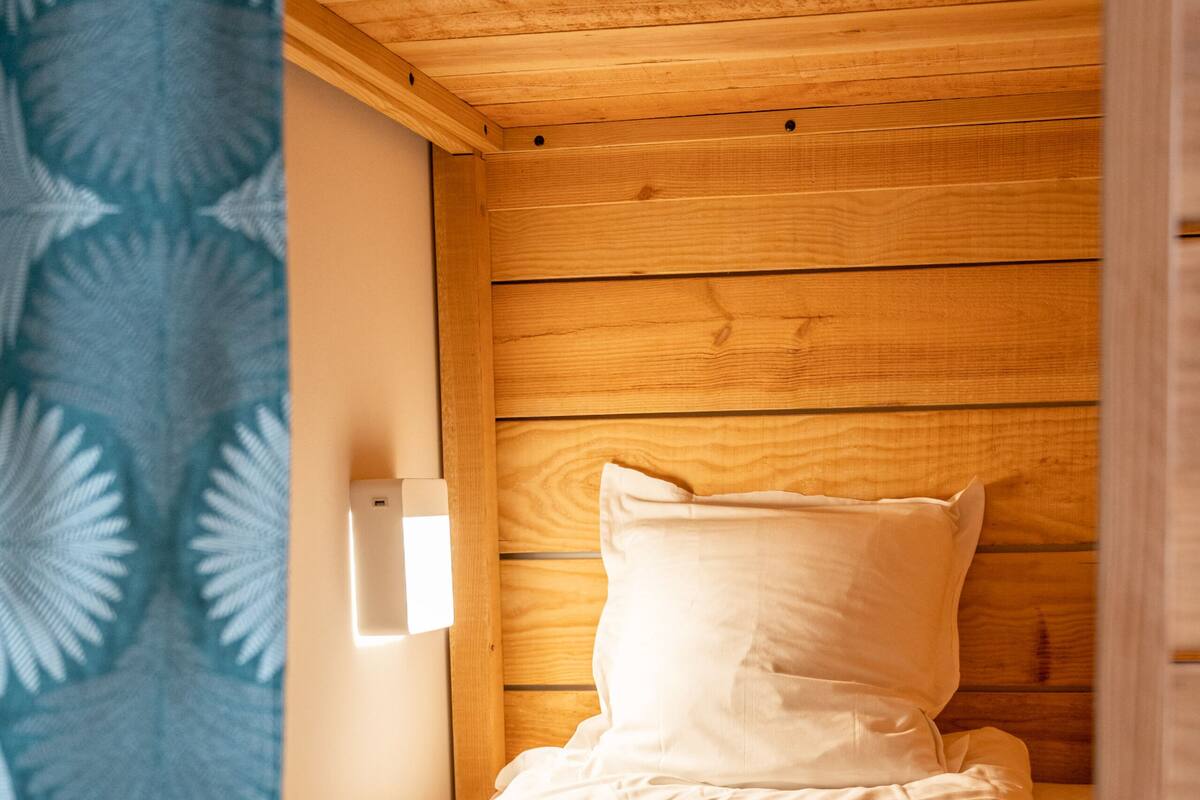 A small wooden room with a bed, a wall-mounted light, and a patterned curtain.