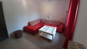 Ein kleiner, gut beleuchteter Raum mit einem roten, modularen Sofa, einem weißen Couchtisch und einem kleinen Hocker. Eine Wandleuchte und eine Stehlampe sind zu sehen.