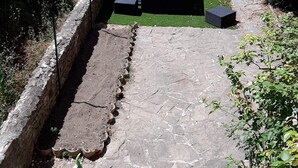 Ein Steinweg führt zu einer Sitzgelegenheit mit einem blauen Sonnenschirm, umgeben von Grünpflanzen und einer Steinmauer.