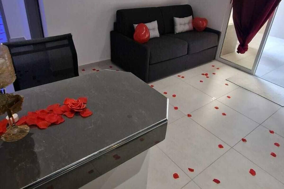 Un salon moderne avec un canapé noir, une table basse en verre et des pétales de rose éparpillés sur le sol.