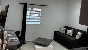 Una sala de estar moderna con un sofá negro, una mesa de centro, un televisor y un ventanal con vistas a edificios.