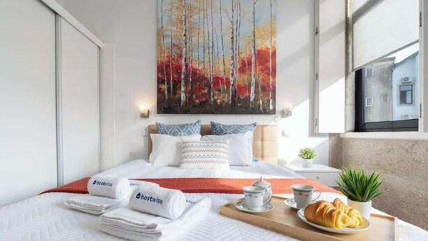 A modern hotel room with a large bed, a painting of trees, a tray with croissants, and a view of a building outside. - Cozy House w Balcony by HostWise (Porto)