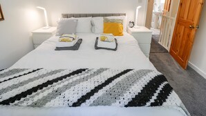 Una cama bien hecha con una colcha a rayas, dos mesitas de noche con lámparas, una puerta de madera y un pasillo que da a un baño.