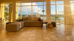 A spacious living room with a large sectional sofa, a small side table, and a view of a lake and mountains through large windows. - Best View-Stylish Penthouse with communal pool (Kerkira)