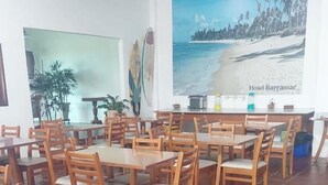 Un comedor con mesas y sillas de madera, un cartel grande con una escena de playa y plantas en macetas.