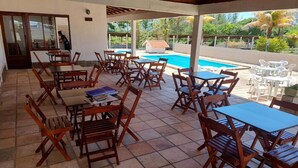 Un espacio al aire libre cubierto con mesas y sillas de madera, una piscina y vistas a la vegetación.