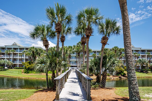 A wooden boardwalk leading to a building with multiple balconies, surrounded by palm trees and a body of water.