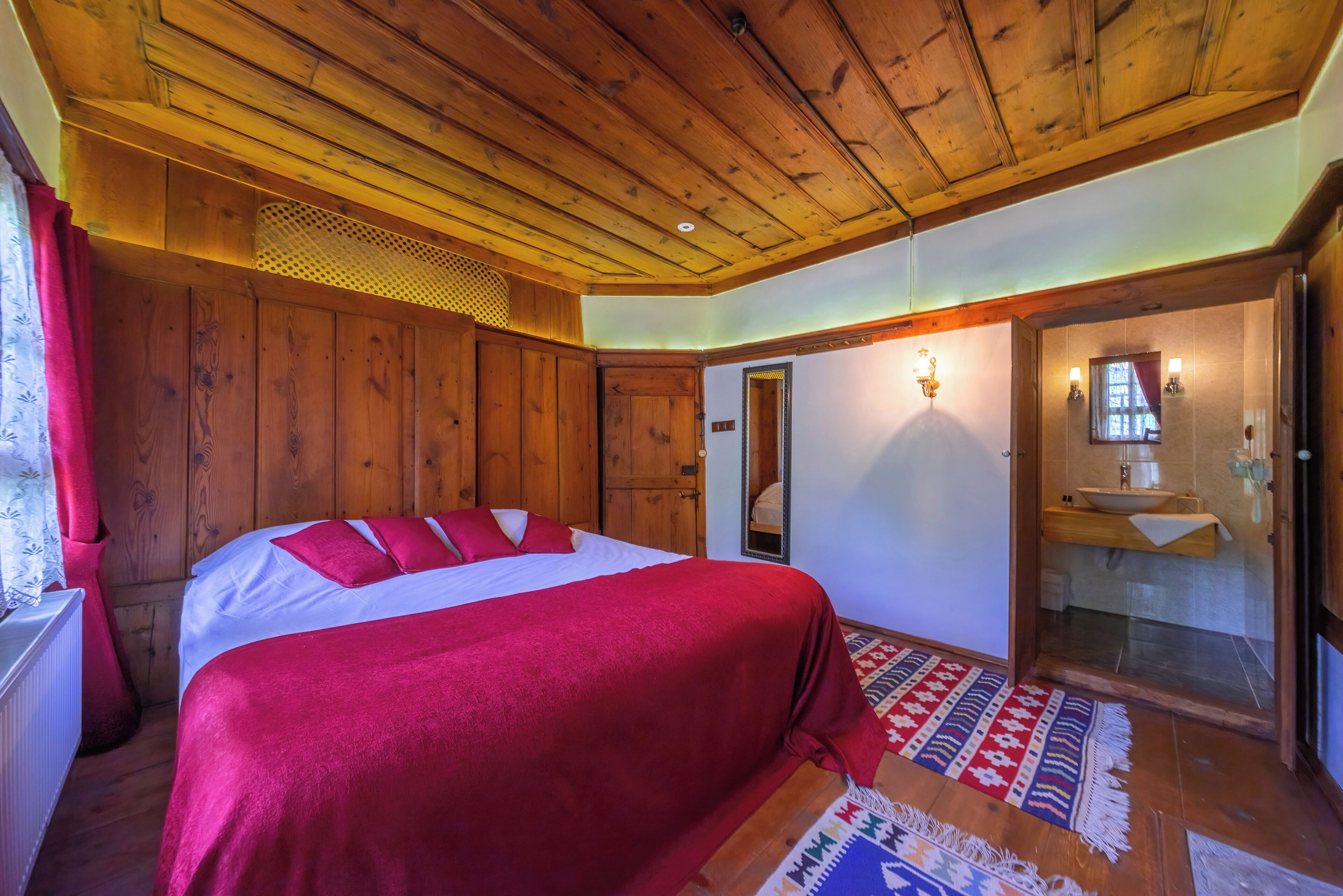 A bedroom with a wooden ceiling, a large bed with red bedding, a mirror, and a bathroom with a sink and towel rack.