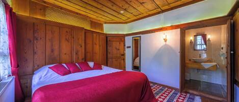 A bedroom with a wooden ceiling, a large bed with red bedding, a mirror, and a bathroom with a sink and towel rack.
