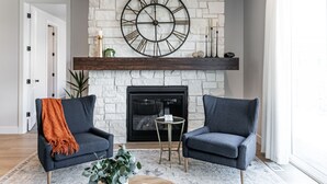 A living room with a fireplace, two armchairs, a round wall clock, and a small table.