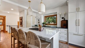 A modern kitchen with white cabinetry, a central island with bar stools, pendant lighting, and a view of the outdoors through a window. - Vintage Modern near Downtown - Arcade Machine (Rapid City)