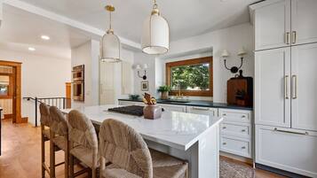Une cuisine moderne avec des meubles blancs, un îlot central doté de tabourets, des luminaires suspendus et une vue sur l’extérieur grâce à une fenêtre.
