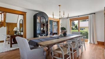 A dining area with a wooden table, white chairs, a chandelier, and a view of greenery outside.