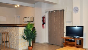 A living room with a wooden coffee table, a TV on a wooden stand, a potted plant, a fire extinguisher, and a kitchen area with a stone wall and a bar counter.