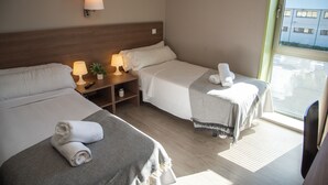 A hotel room with two beds, a nightstand with lamps, a small shelf with a plant, and a window with blinds. - Hostal La Parada (Getafe)