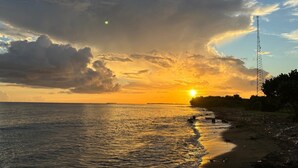 A sunset over a beach with a clear sky, a water body, and a distant landmass. - The Place at the Sea (Ponce)