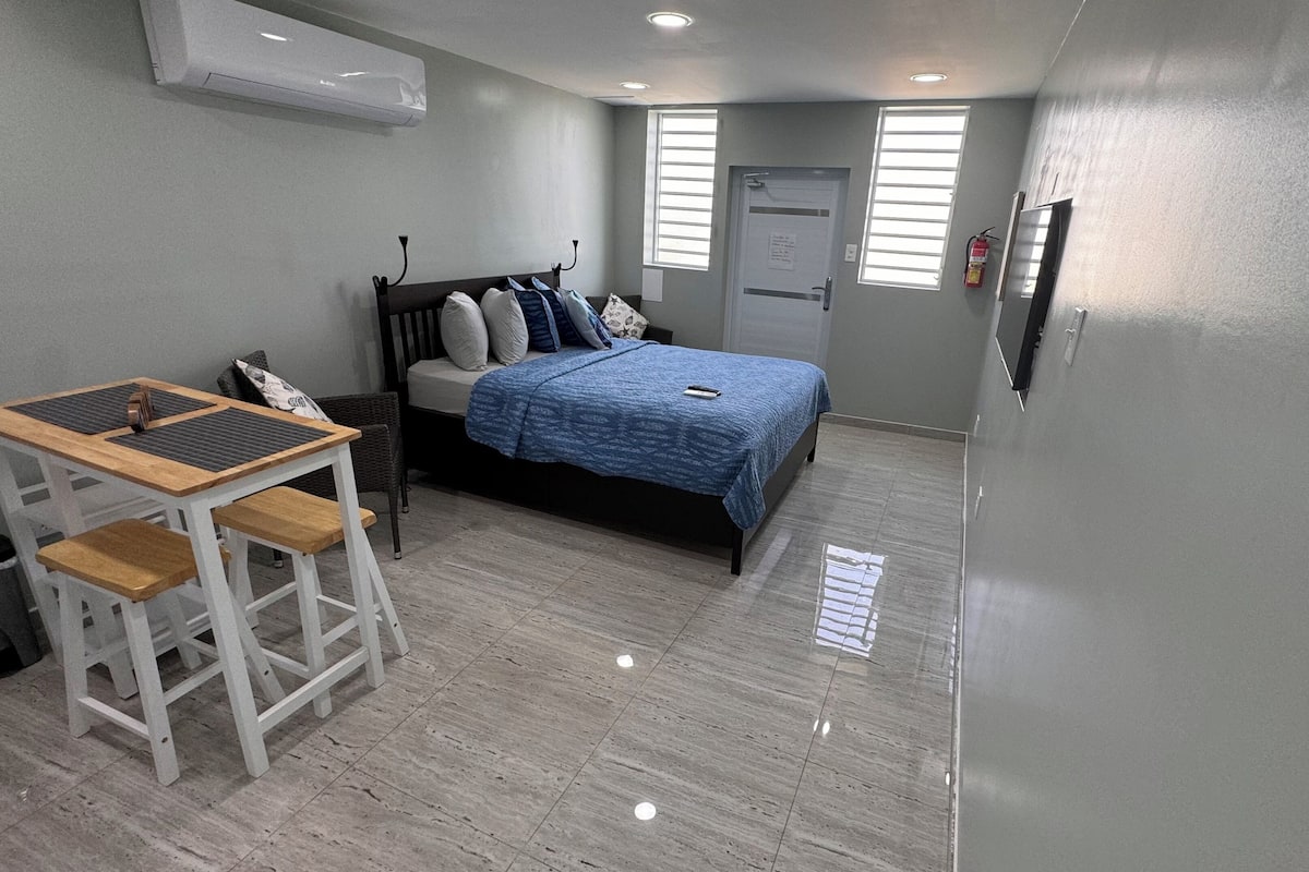A bedroom with a bed, a desk, and a chair. There is a window with blinds, a door, and a fire extinguisher on the wall.