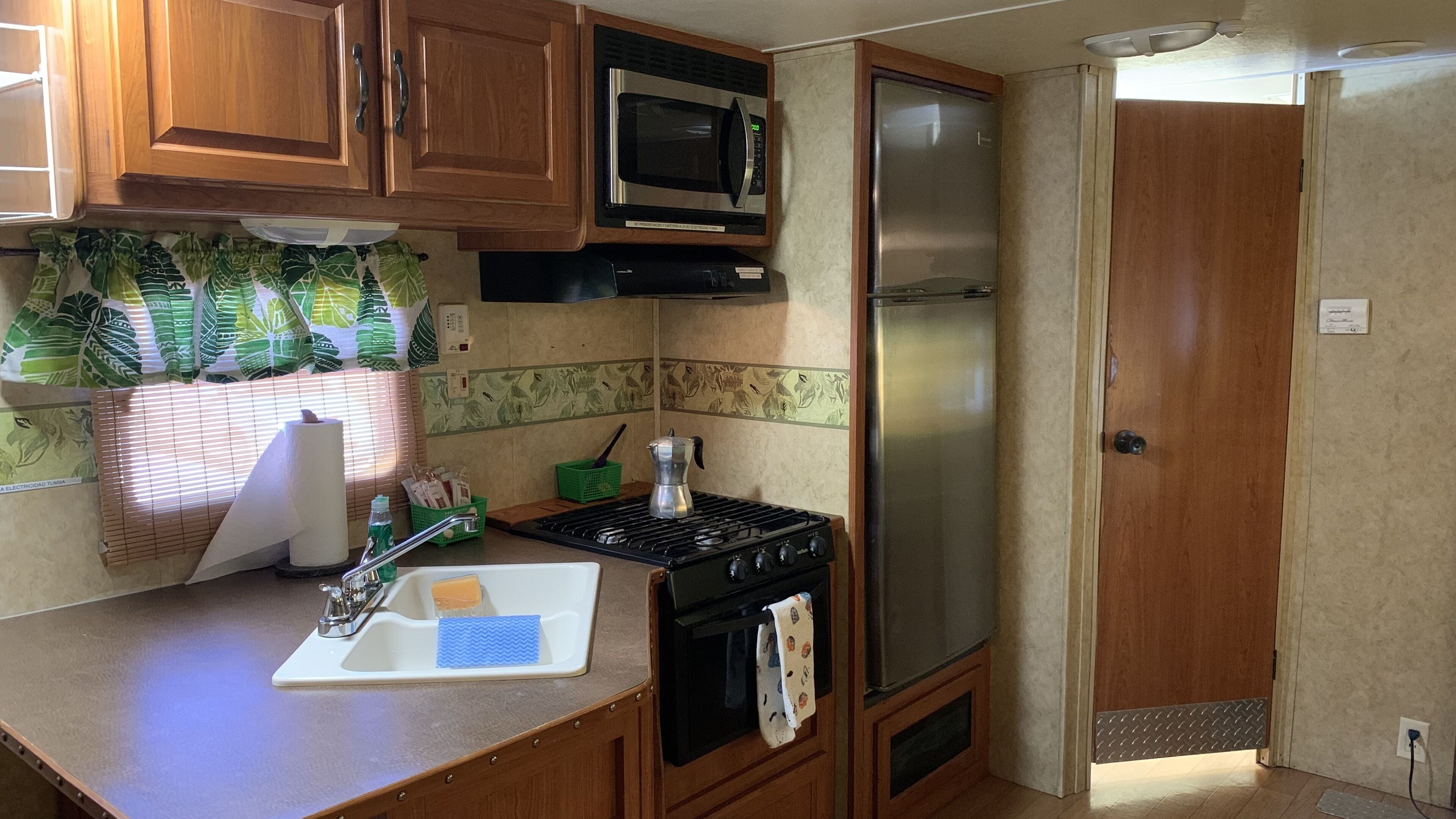Una cocina compacta con armarios de madera, un microondas, un refrigerador de acero inoxidable, una estufa y un lavamanos con un jabonera.