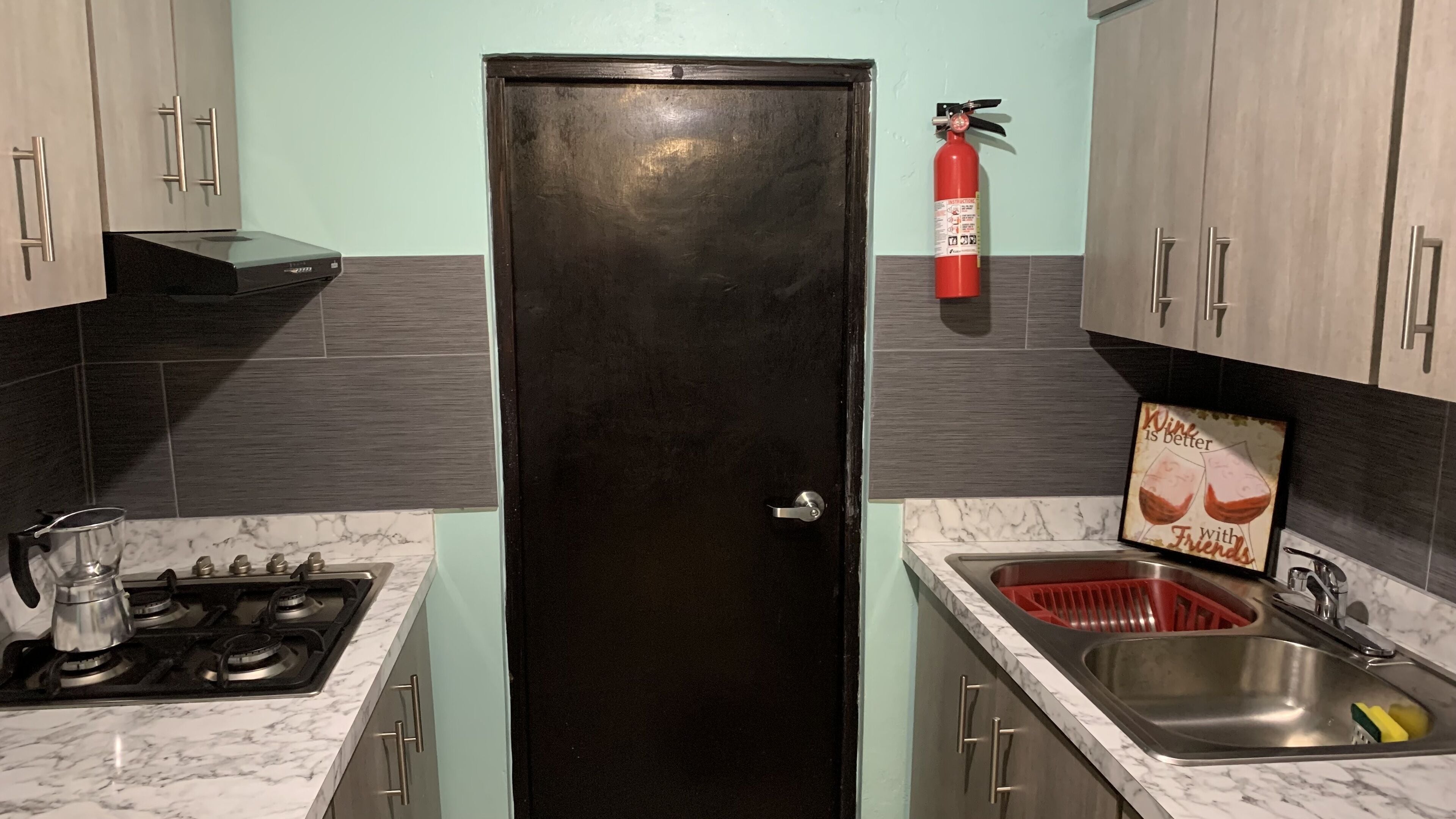 A compact kitchen with a black refrigerator, a gas stove, a sink with a red dish rack, and a fire extinguisher mounted on the wall.