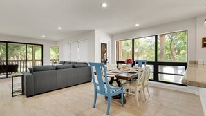 A spacious living area with a grey sofa, a dining table with chairs, and large windows offering a view of greenery.