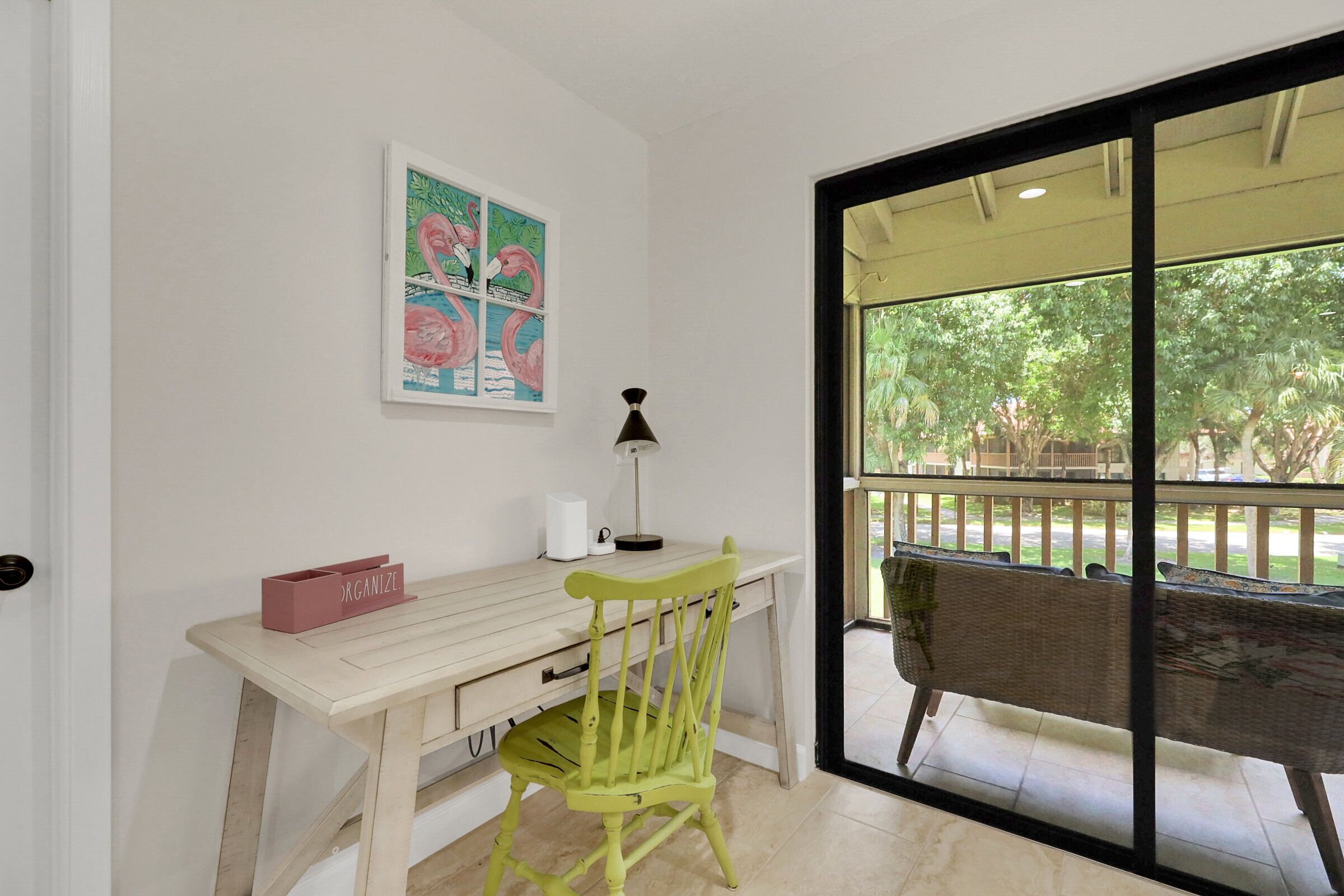 A room with a desk, chair, and a window with a view of trees and a balcony.