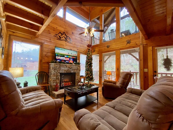 A cozy cabin interior with a stone fireplace, comfortable seating, a TV mounted on the wall, and a Christmas tree. - ✨Luxury Creekside Cabin, No Steep Roads, Pet Friendly, Hot Tub, Family Games✨ (Pigeon Forge)
