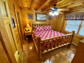 A wooden cabin bedroom with a bed, a nightstand, a ceiling fan, and framed pictures on the walls. - ✨Luxury Creekside Cabin, No Steep Roads, Pet Friendly, Hot Tub, Family Games✨ (Pigeon Forge)