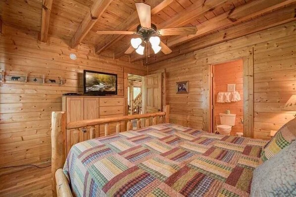 A wooden cabin bedroom with a bed, a ceiling fan, a television, and a wooden dresser.