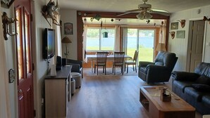 Un salon chaleureux avec un canapé en cuir noir, une table basse en bois et un téléviseur. On y trouve également un coin repas avec une table et des chaises, ainsi qu’une grande fenêtre donnant sur l’extérieur.